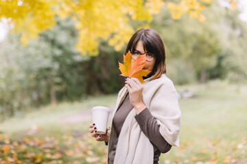 Playful girl in glasses hold in hand white mock up cup with tea or coffee and hiding behind orange maple leaf. Woman looking into camera.  Hot warming beverage. Autumn season. Blurred background.