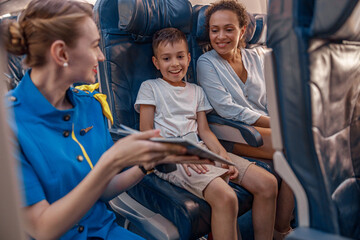 Wall Mural - Female air hostess trying to entertain a kid on the plane by offering a book to read. Cabin crew provide service to family in airplane. Airline transportation and tourism concept