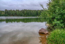 Summer Landscape On The Shore Of A Forest Lake On A Cloudy Day. In The Foreground, Among The Bushes Of Greenery, Lies A Large Stone. The Forest And Hills Are Visible In The Distance. Russia (Ural)
