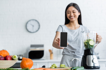 Wall Mural - blurred asian woman showing cellphone with blank screen near blender and fresh vegetables.