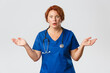 Worried and confused redhead female doctor, nurse cant understand what happened, wearing blue scrubs, spread hands sideways and looking concerned and nervous at camera, grey background