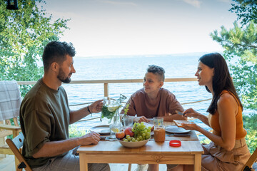 Happy family sitting by served table during outdoor dinner or breakfast against waterside