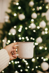 Wall Mural - Stylish cup in hand on background of christmas tree in lights in evening room. Cozy winter time. Space for text. Female hand in sweater holding ceramic cup with tea in festive atmospheric room