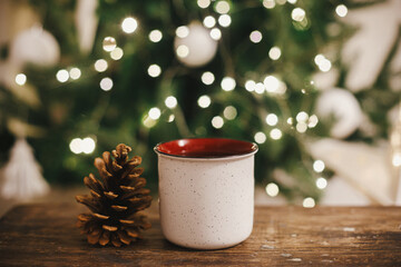 Stylish cup and pine cone on rustic wood on background of christmas tree in lights in evening room. Space for text. Ceramic cup with tea in festive decorated room. Atmospheric christmas time