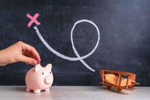 A Woman's Hand Puts A Coin In A Piggy Bank Standing Next To A Wooden Airplane A Toy On A Chalk Board Background