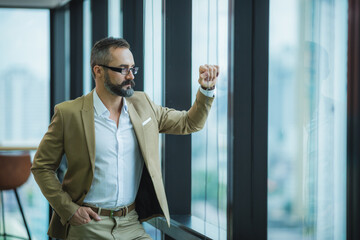 Wall Mural - businessman look at the office's window, relax and chill moment during office time, modern lifestyle, smart business man in light brown outfits, caucasian man, city view background.