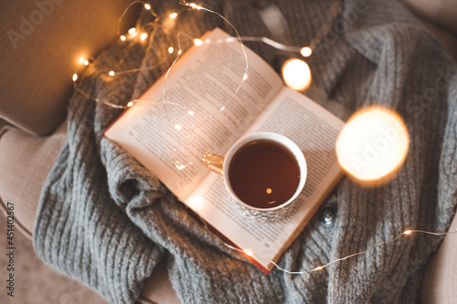 Cup of fresh black tea stay on open paper book with knit woolen sweater in cozy chair over glow Christmas lights closeup. Top view. Early morning breakfast. Winter comfy home atmosphere.