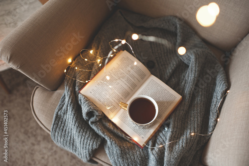 Cup of fresh black tea stay on open paper book with knit woolen sweater in cozy chair over glow Christmas lights closeup. Top view. Early morning breakfast. Winter comfy home atmosphere.