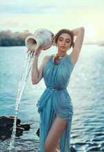 Art Photo. Portrait Mythical Girl Greek Goddess. Astrological Sign Aquarius. Fantasy Woman Pours Water From Ceramic Clay Jug. Blue Chiffon Old Vintage Long Silk Dress. Fabulous Lake, Magical Sunset
