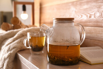 Poster - Glass teapot and cup of hot tea near open book on wooden table in room. Cozy home atmosphere