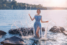 Greek Mythical Fairytale Fantasy Woman Goddess Nymph Emerges From Lake. Splashes Of Water. Vintage Blue Long Sexy Wet Dress. Summer Nature Forest, Water Surface Of River Coastal Stones. Girl Mermaid