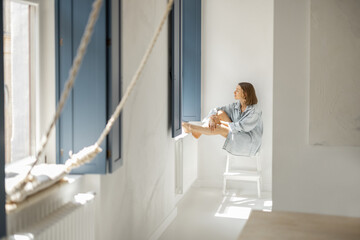 Wall Mural - Young woman sitting alone by the window in a bright room at home. Self-isolation, loneliness, girl feeling calm and relaxed at home