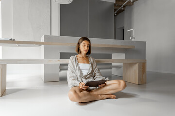 Canvas Print - Young woman works on a digital tablet while sitting relaxed at modern kitchen interior. Remote work from home, comfortable and modern apartment