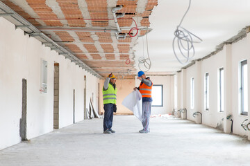 Two men with plan wearing safety vests talking in building under construction