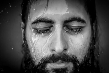 Black and white very close-up portrait of a young bearded man with closed eyes with streams of water running down his face