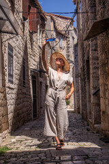 Beautiful blonde young female traveler wearing straw sun hat, taking selfie while sightseeing and enjoying summer vacation in an old traditional costal town at Adriatic cost, Croatia.