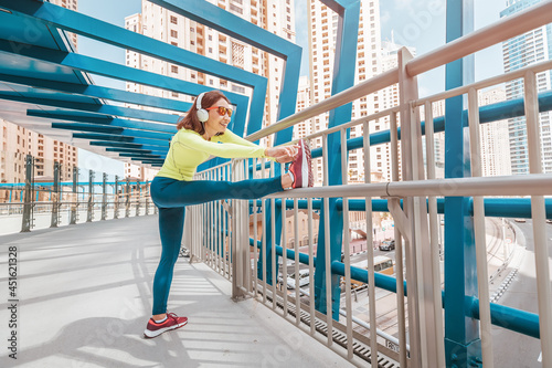 Happy woman wearing headphones stretches the quadriceps muscles of the thigh and warms up before intense running workout on the foot brige in Dubai Marina