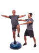 An elderly man doing a balance exercise, on one leg, on a hemisphere ball. With help of a fitness trainer. On a white isolated background.