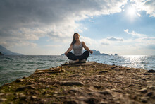 Young Active Brunette Is Sitting In Lotus Position On Rocky Seashore, Front View From Low Angle, Beautiful Seascape On Background. Woman On Fitness And Yoga Tour For Reboot.