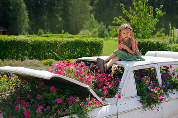 A girl with African pink pigtails from kanekalon is sitting on a designer landscape car with flowers. A flower garden of petunias in the garden. Funny kid. A flowerbed with petunia flowers. zizi pigta