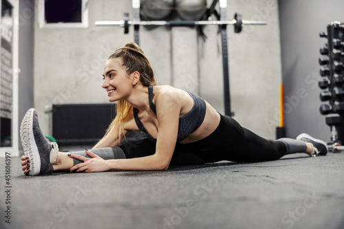 Muscle flexibility and hip mobility. Fit woman in sportswear and good body shape stretches legs muscles on the floor of an isolated gym. Enjoyment and smile in training, ponytail