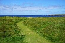 Green Grass Pathway Leading Across Overgrown Moors To The Bright Blue Sea Under A Lightly Cloudy Sky In Rural Scotland