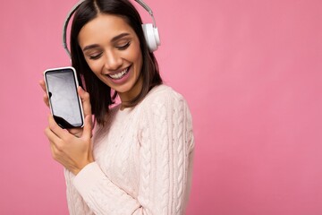 Wall Mural - Pretty young brunette woman wearing pink casual sweater isolated over pink background wall wearing white bluetooth wireless headphones and listening to good music and showing mobile phone with empty