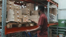 Slowmo Tracking Shot Of African-American Male Warehouse Worker Using Barcode Scanner And Checking Cardboard Boxes Stacked On Shelf