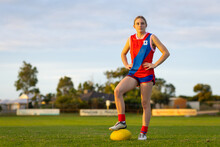 young female aussie rules footballer standing with hands on hips and foot on ball