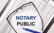On a light background, a report, black-framed glasses, a pen and a sheet of paper with the text NOTARY PUBLIC. Business concept
