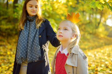 Wall Mural - Two cute young sisters having fun on beautiful autumn day. Happy children playing in autumn park. Kids gathering yellow fall foliage.