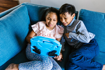 Happy latin siblings sitting on sofa using digital device at home in Mexico