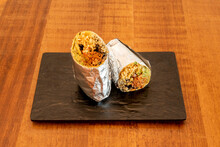 Shredded Beef Burrito With Lettuce And Black Beans, White Rice And Wheat Tortilla Wrapped In Aluminum Foil On A Black Slate Tray