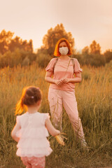 Young redhead woman walking with cute baby girl in  summer meadow. Girl wearing face mask outside.