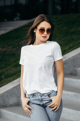 Stylish brunette girl wearing white t-shirt and glasses posing against street , urban clothing style. Street photography