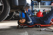 © Wavebreak Media - Mixed race female car mechanic wearing overalls, lying on a board, checking car