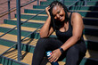African Exhausted Female In Black Sportive Clothes Have Rest After Sport, Sitting With Water Bottle, Having Rest, Looking At Camera Smiling, Posing. In The Morning, Weight Loss, Sport, Fitness