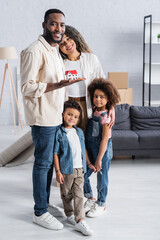 happy african american man holding house model near wife and children in new apartment