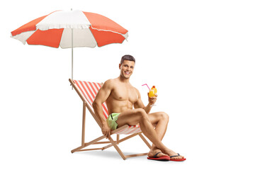 Wall Mural - Guy in swimming shorts sitting on a beach chair under umbrella and holding a cocktail