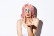 Close-up of lovely girl in halloween costume with pink wig and bright makeup, close eyes and blowing air kiss at camera, standing over white background