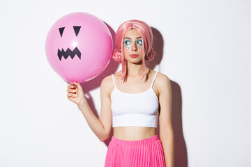 Image of beautiful glamour girl in pink wig, bright makeup, holding balloon with scary halloween face, standing over white bacground