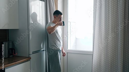 Handsome young man pours coffee from coffee maker and walks up to window in kitchen.