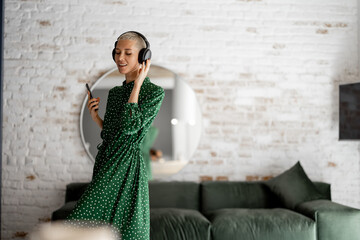 Wall Mural - Stylish woman in green dress enjoys the music with headphones and cell phone dancing in the living room at home. Comfort living and leisure time concept