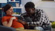 © DC Studio - Pregnant interracial couple sitting on living room couch. African american husband touching baby bump and talking to child while caucasian mother relaxing on sofa and laughing