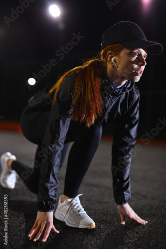 Fitness woman runner in start position outdoors at night in city. Woman running, jogging. confident woman standing low start before running. Concept of jogging, training outdoor, active lifestyle.