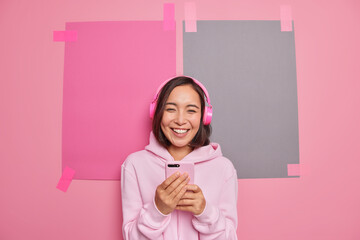 Wall Mural - Happy young Asian woman holds smartphone wears stereo headphones on ears listens music from playlist wears hoodie poses against pink background with plastered sheets to place your information