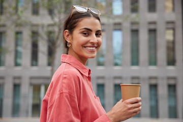 Positive dark haired woman in stylish red shirt sunglasses on head strolls in unknown city drinks coffee from paper cup enjoys leisure time smiles broadly poses against blurred building background