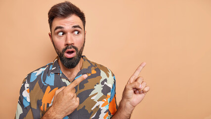 Wall Mural - Shocked bearded man shows unexpected revelation indicates at promo shows copy space wears colorful shirt isolated over beige background gasps overwhelmed stares surprised. Omg look at this advert