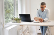 © epiximages - business man with blue shirt and black glasses is standing behind standing table and is working with his tablet and a black laptop in a modern office