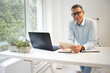 © epiximages - business man with blue shirt and black glasses is standing behind standing table and is working with his tablet and a black laptop in a modern office